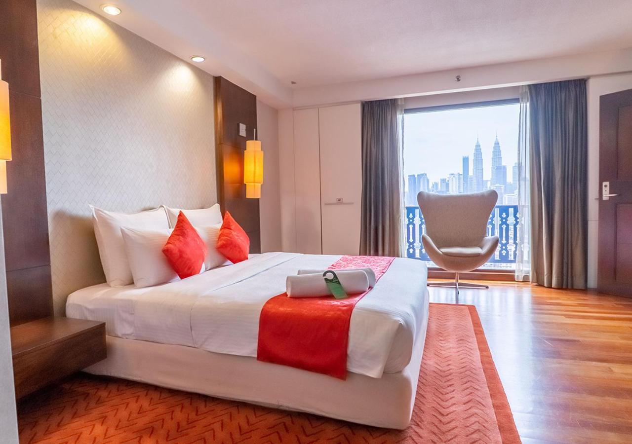 Seri Pacific Hotel Куала-Лумпур Экстерьер фото The photo shows a modern hotel room featuring a neatly made bed with white linens and red accent cushions. There is a stylish orange rug on the floor, and a large window offers a view of a city skyline, including tall buildings. A comfortable-looking