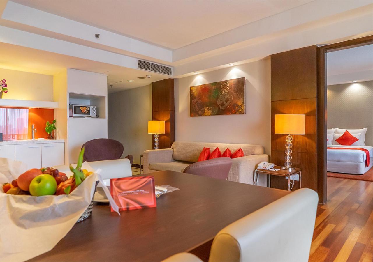 Seri Pacific Hotel Куала-Лумпур Экстерьер фото The photo shows a well-decorated hotel suite or living area. It features a cozy seating arrangement with a sofa and chairs, accompanied by bright throw pillows. There's a dining table in the foreground topped with a fruit basket. The room has warm li