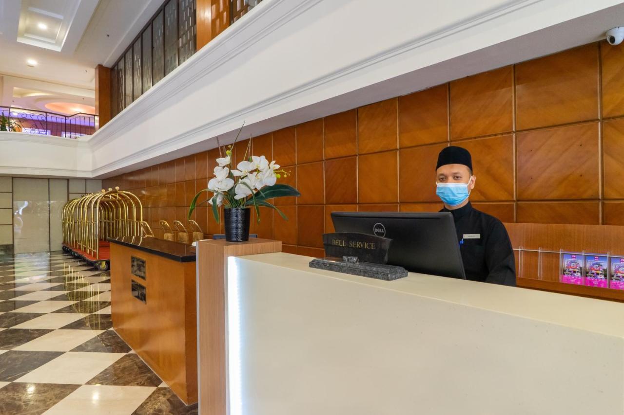 Seri Pacific Hotel Куала-Лумпур Экстерьер фото The photo shows a hotel reception area. In the foreground, a staff member is standing behind a reception desk, wearing a mask and a dark uniform. The desk has a computer monitor, and there are decorative elements, including a vase with flowers. Behin
