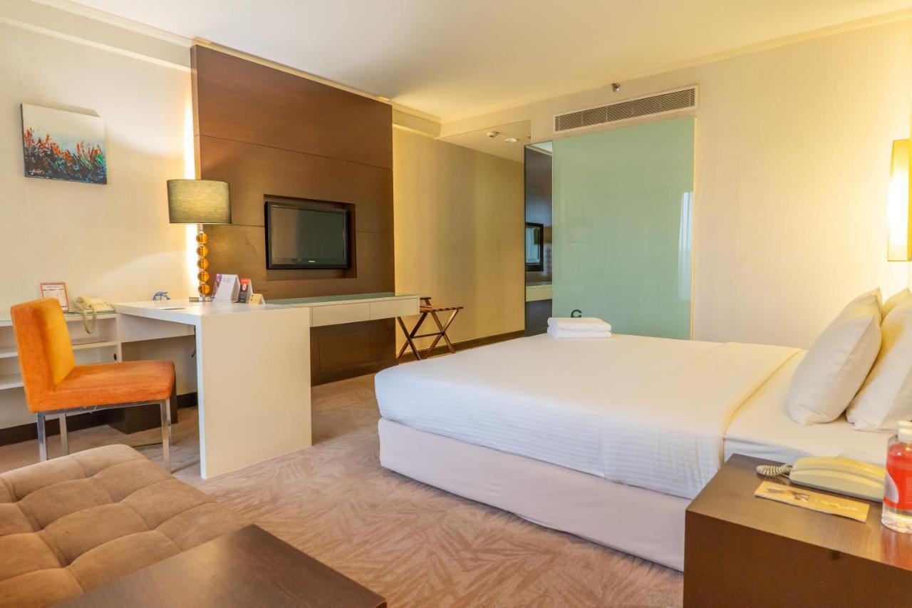 Seri Pacific Hotel Куала-Лумпур Экстерьер фото The photo shows a hotel room or a similar accommodation space. It features a large bed with white bedding, a bedside table with a lamp, and a small seating area. There is a desk with a chair, and a television mounted on the wall. A glass partition se