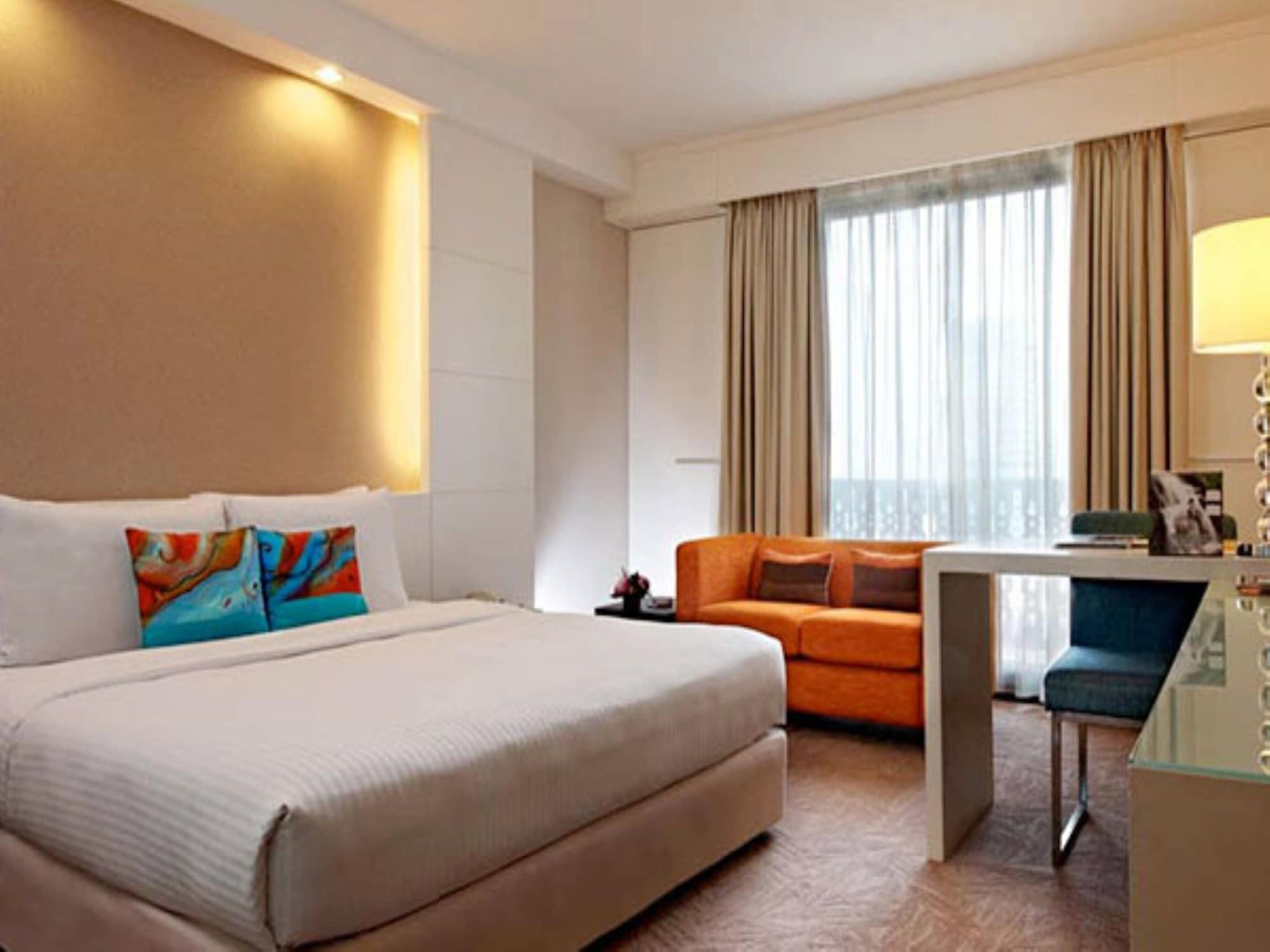 Seri Pacific Hotel Куала-Лумпур Экстерьер фото The photo shows a modern hotel room featuring a large bed with white bedding. There are colorful accented pillows on the bed. To one side, there is an orange sofa that adds a pop of color to the space. A large window with light curtains allows natura