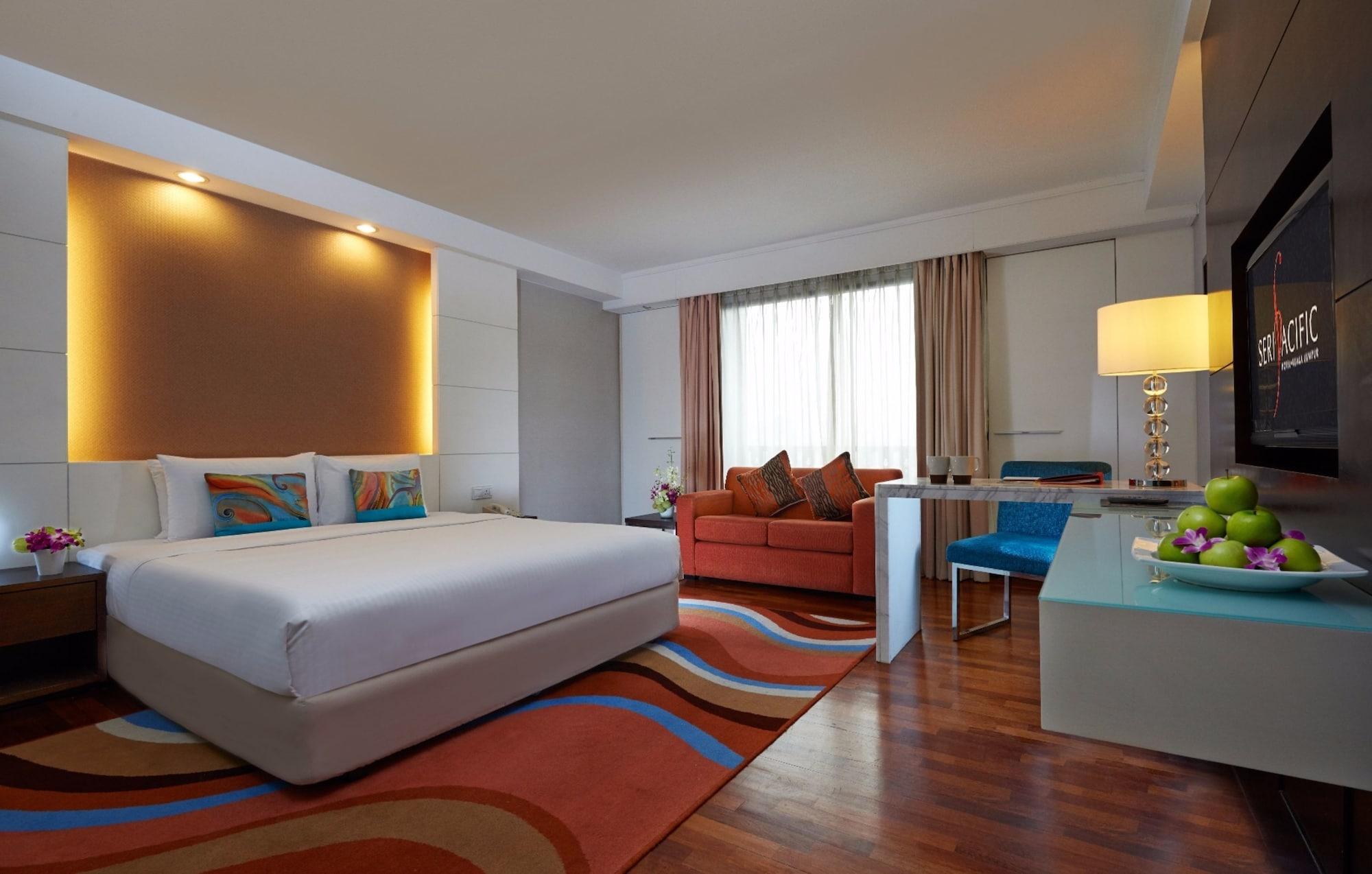 Seri Pacific Hotel Куала-Лумпур Экстерьер фото The image shows a well-decorated hotel room. On one side, there is a large bed with a light-colored headboard, complemented by colorful pillows. A cozy orange sofa is positioned across from the bed, creating a comfortable seating area. There is a mod