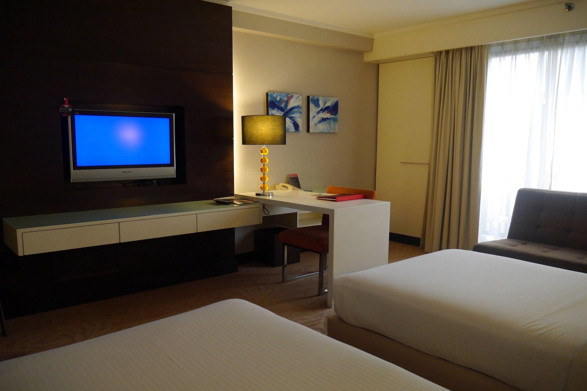 Seri Pacific Hotel Куала-Лумпур Экстерьер фото The photo shows a modern hotel room featuring two beds with white linens. There is a flat-screen TV mounted on the wall in front of the beds. Next to the TV, there is a desk and a chair, along with a lamp. The room has light-colored walls and curtain