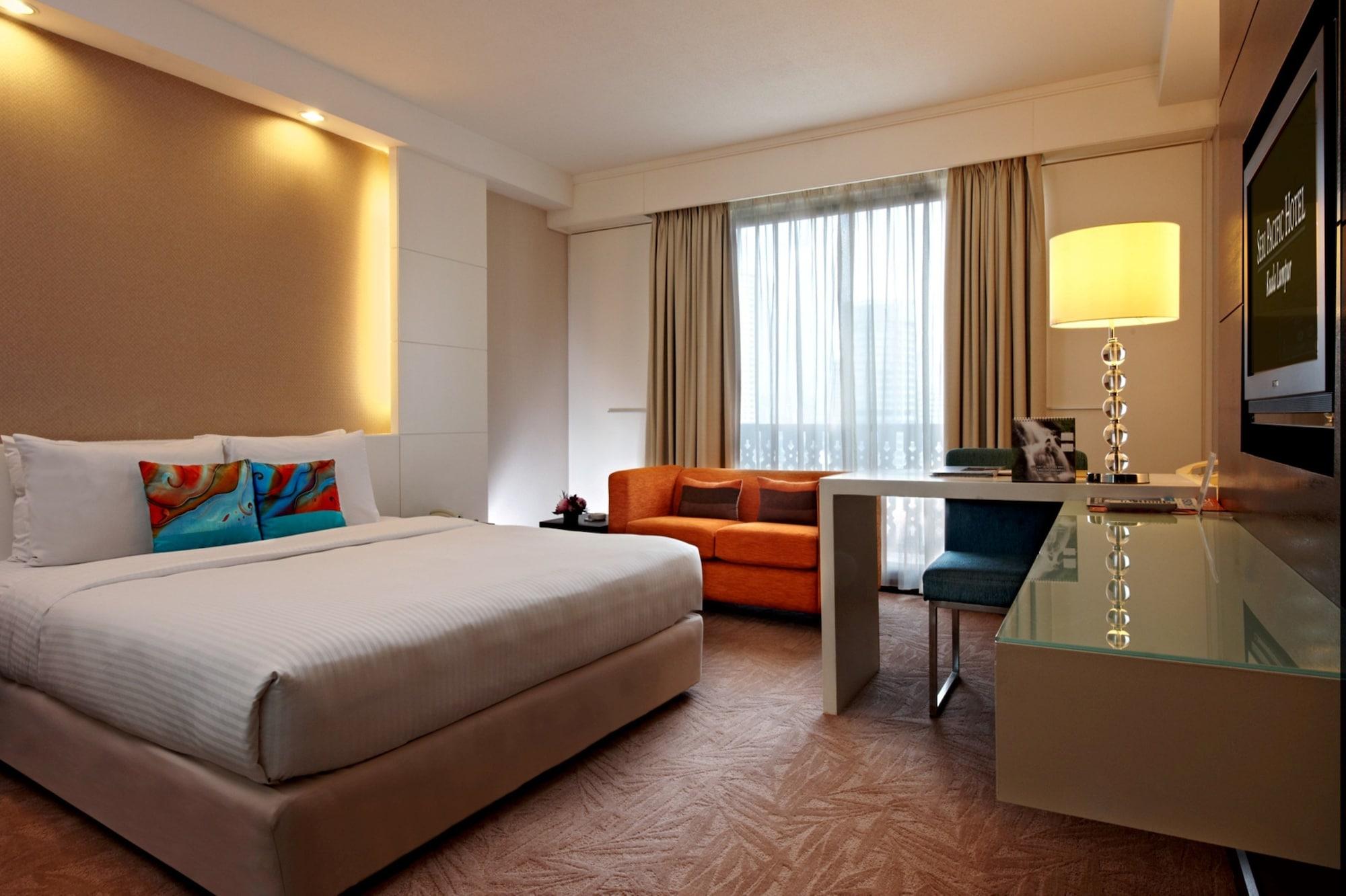 Seri Pacific Hotel Куала-Лумпур Экстерьер фото The photo shows a modern hotel room. It features a large, neatly made bed with decorative pillows at the head. There is a small sofa in a vibrant orange color next to the bed. A large window with sheer curtains lets in natural light, and there's a la