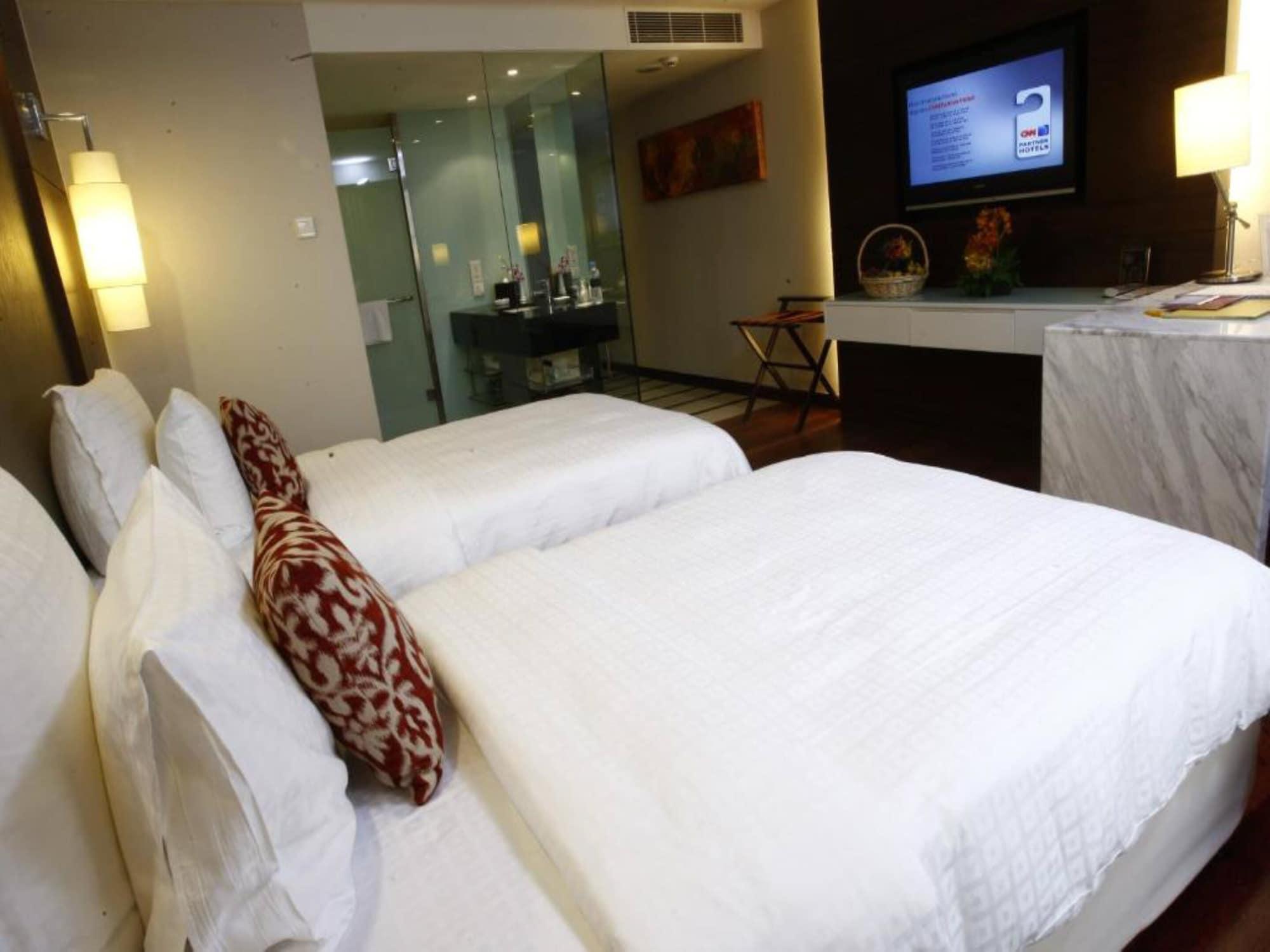 Seri Pacific Hotel Куала-Лумпур Экстерьер фото The photo shows a modern hotel room featuring two twin beds with white bedding and decorative pillows. In the background, there's a glass-walled bathroom visible. There's a flat-screen TV mounted on the wall, and a marble console table or dresser nea