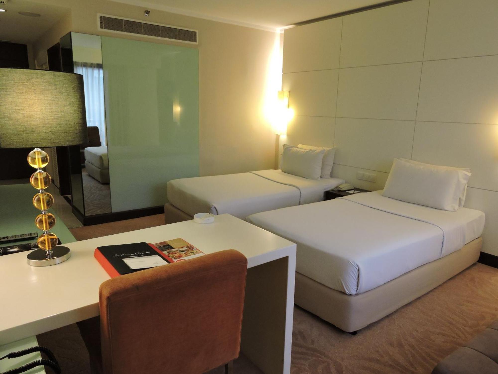 Seri Pacific Hotel Куала-Лумпур Номер фото The photo shows a modern hotel room featuring two single beds with white bedding. There is a minimalist design with a light-colored scheme. A small desk with a brown chair is positioned nearby, along with a lamp that has a decorative base. The room h