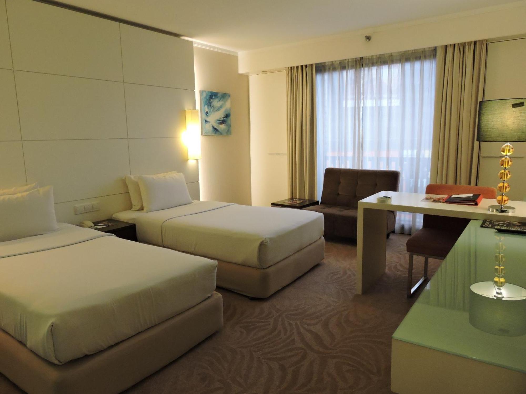 Seri Pacific Hotel Куала-Лумпур Номер фото The photo shows a modern hotel room featuring two twin beds with white bedding. The walls are light-colored with a sleek and minimalist design. There is a large window covered by sheer curtains, allowing natural light to enter the room. To one side, 