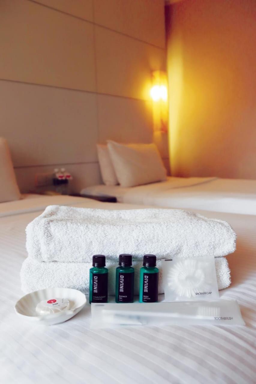 Seri Pacific Hotel Куала-Лумпур Экстерьер фото The photo shows a neatly arranged stack of white towels placed on a bed. In front of the towels, there are three small green bottles, likely containing shampoo, conditioner, and body wash. The background features a well-decorated hotel room with two 