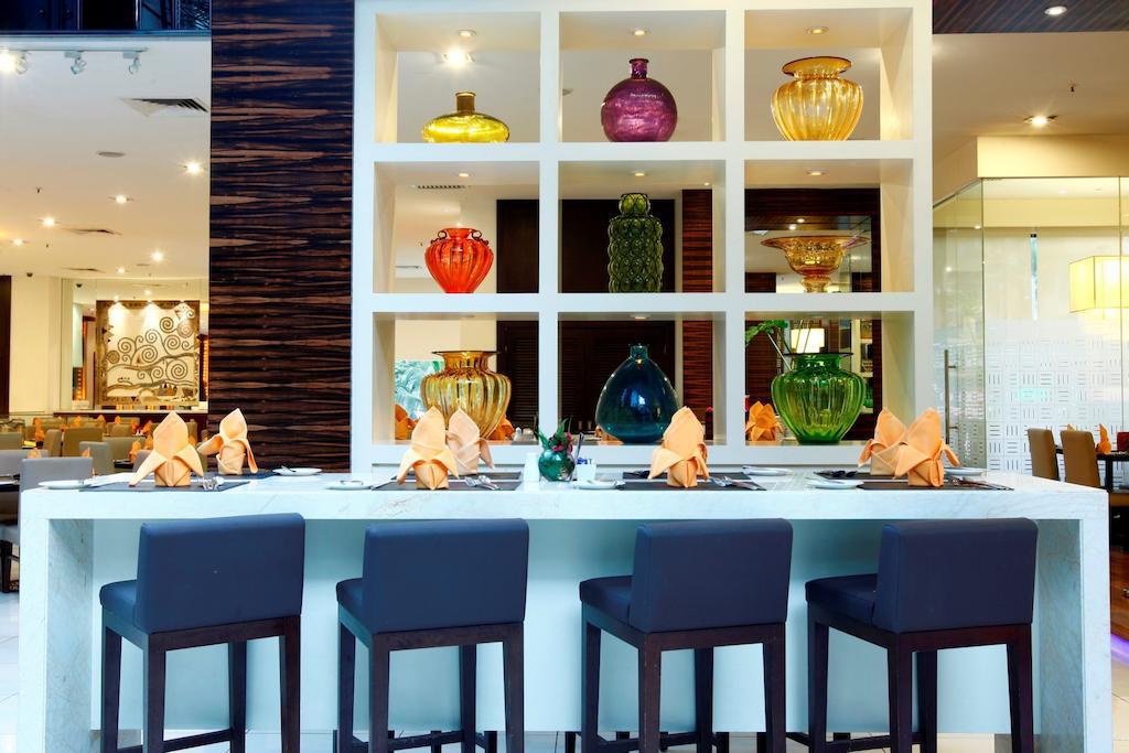 Seri Pacific Hotel Куала-Лумпур Экстерьер фото The photo depicts a stylish restaurant interior featuring a long counter with several dark chairs in front. Behind the counter, there is a large shelf with compartments displaying various colorful glass vases in shades of green, yellow, red, and purp